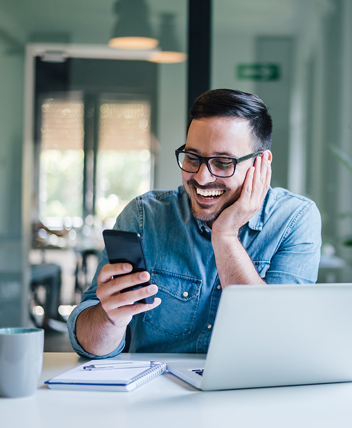 Smiling solopreneur engaging with smartphone while working on laptop, embodying the stress-free financial solutions offered by Mark Rivera, CPA for solo entrepreneurs.